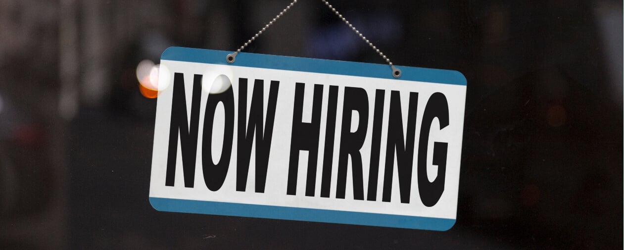 Close-up on a sign in the window of a shop displaying the message: Now hiring