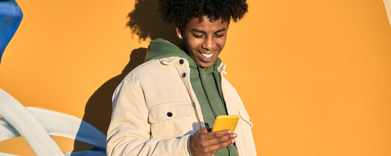 A Get Z young man smiling while holding a phone standing against a brightly colored wall
