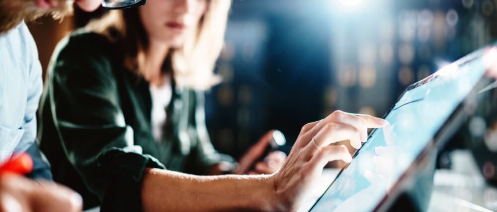 Woman pointing on digital tablet screen while colleague is documenting information on paper.